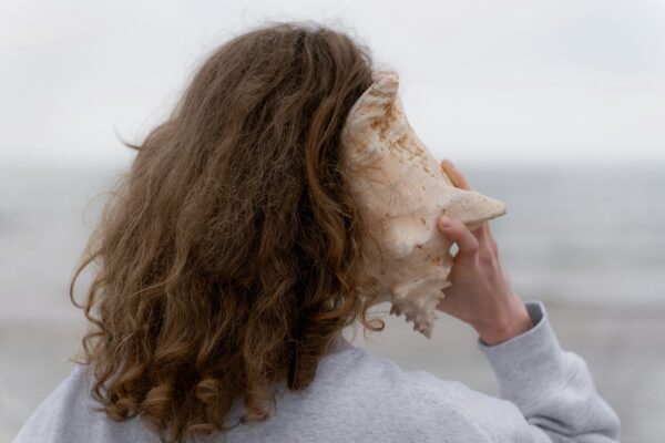 A close up photo of the back of a woman's head. The woman has shoulder length naturally cury hair and she has a large conch shell held up to her ear, she is listening to the sound of the sea