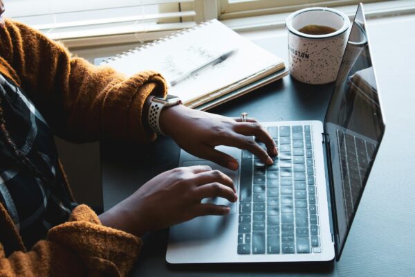 Close up image of a person typing on a laptop keyboard. There is a pen and paper, and a cup of tea, in the background
